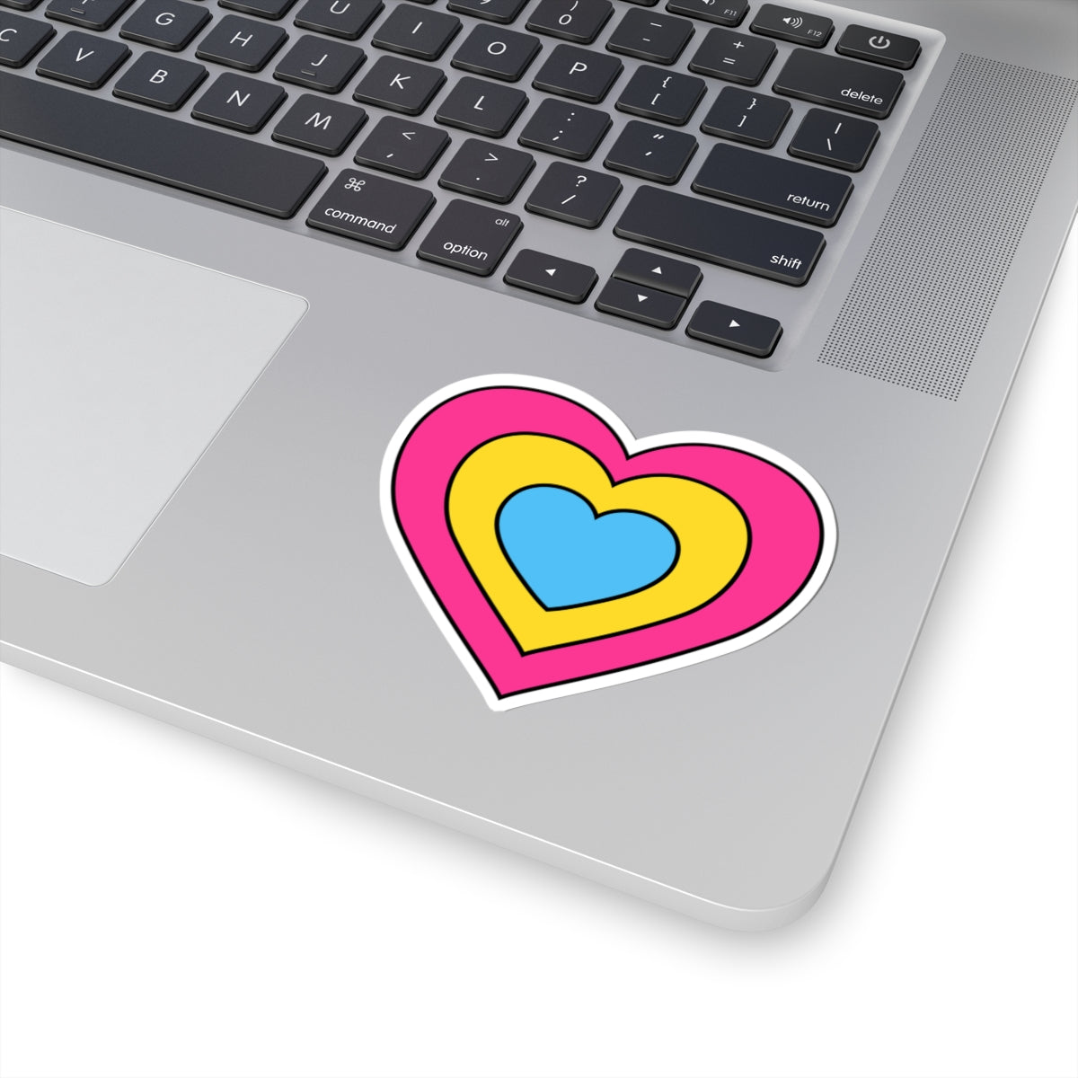 Close-up of the bottom right corner of a silver laptop featuring a 3 inch by 3 inch pride flag heart sticker in a Pansexual flag style applied to the surface.